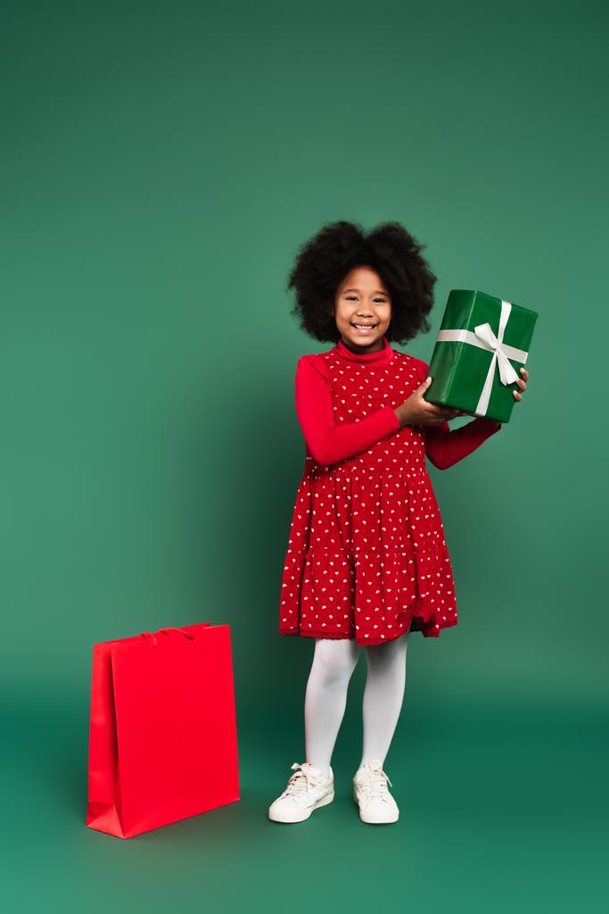Щаслива африканська дитина, яка тримає подарункову коробку з стрічкою біля сумки для покупок на зеленому тлі.  - Фото, зображення