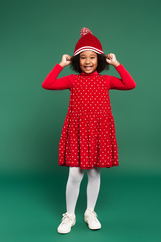 Полная длина улыбающейся африканской девушки в красном платье в шляпе на зеленом фоне  - Фото, изображение