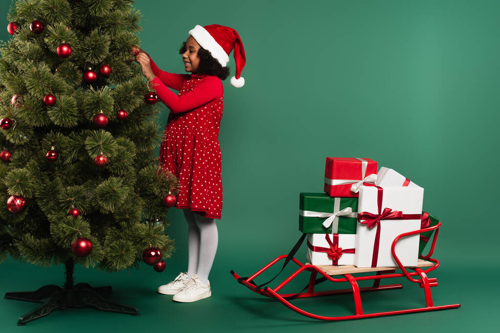Vue latérale de souriante fille afro-américaine en santa chapeau décorer arbre de Noël près du traîneau avec des cadeaux sur fond vert  - Photo, image