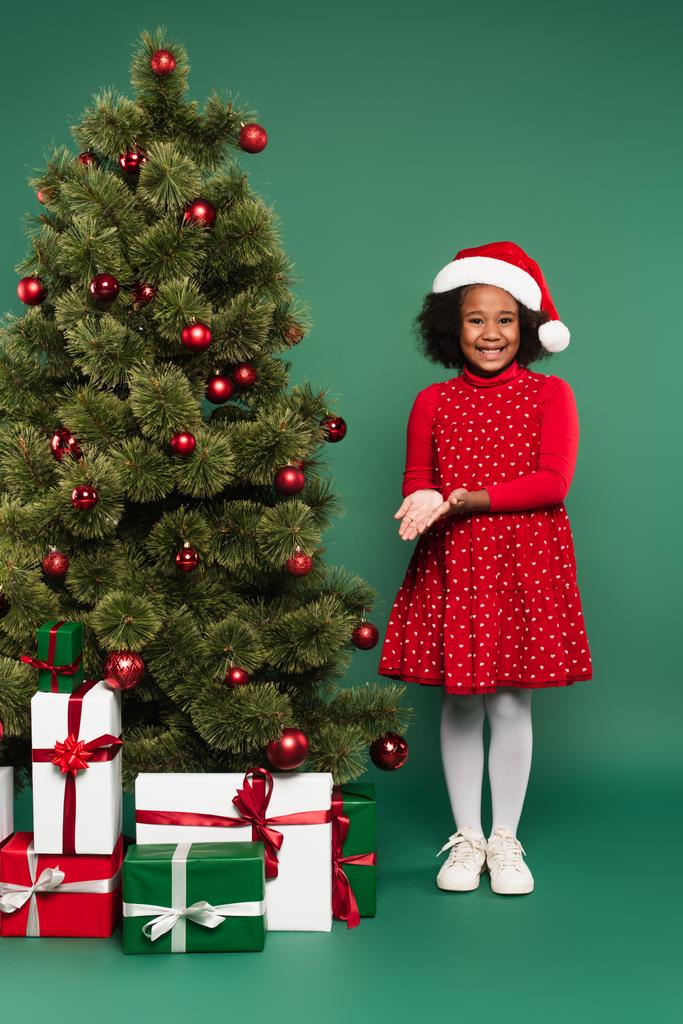 glimlachend Afrikaans amerikaans meisje in santa hoed wijzend op geschenken onder kerstboom op groene achtergrond  - Foto, afbeelding