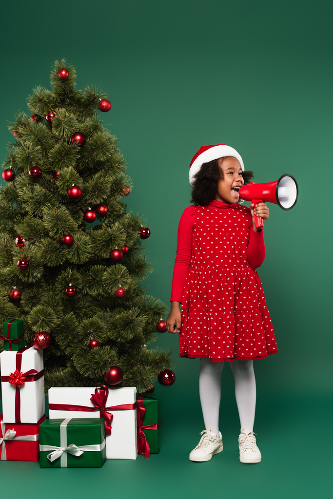 Afroamerikanerin mit Weihnachtsmütze spricht im Lautsprecher neben Weihnachtsbaum und Geschenken auf grünem Hintergrund  - Foto, Bild