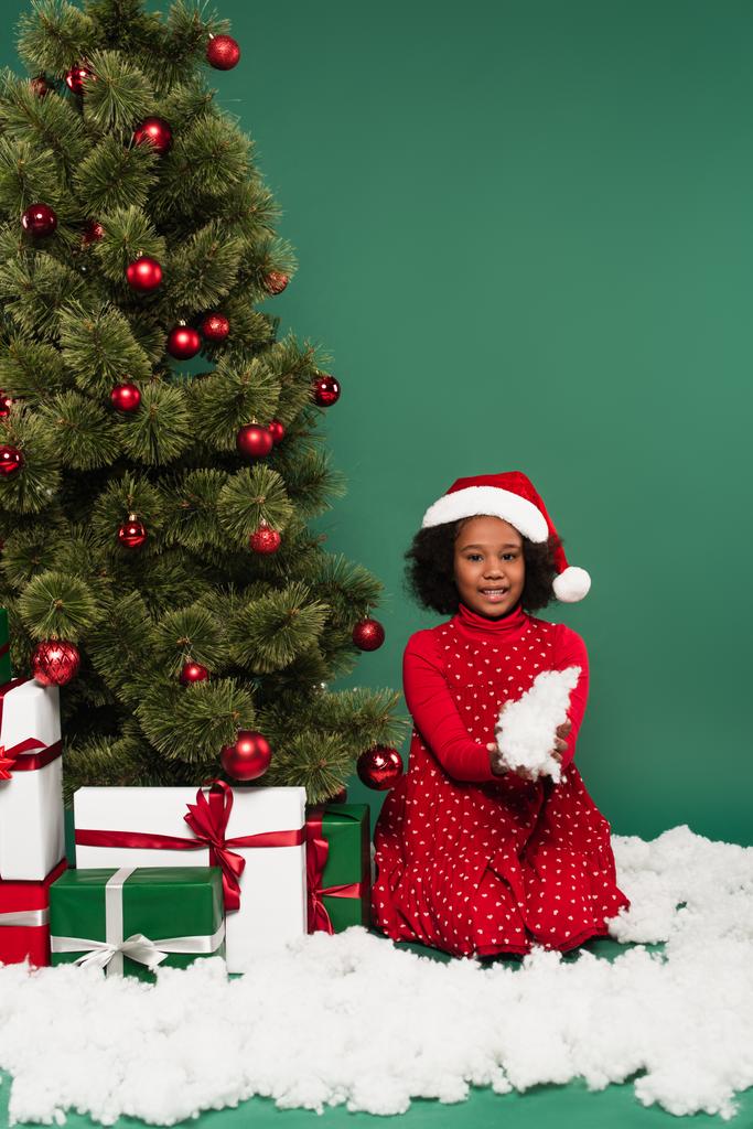 Afrikaans amerikaans kind met decoratieve sneeuw in de buurt van cadeautjes en kerstboom op groene achtergrond  - Foto, afbeelding