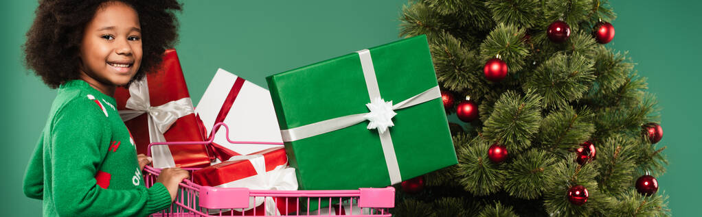 Lächelndes afrikanisch-amerikanisches Kind im Pullover neben Einkaufswagen mit Geschenken am Weihnachtsbaum isoliert auf grünem Banner  - Foto, Bild