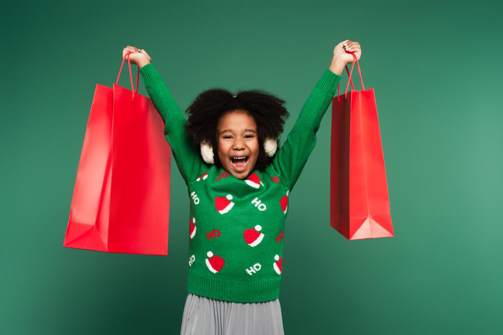 Podekscytowana Afroamerykanka w swetrze i nausznikach trzymająca czerwone torby na zakupy odizolowane na zielono - Zdjęcie, obraz