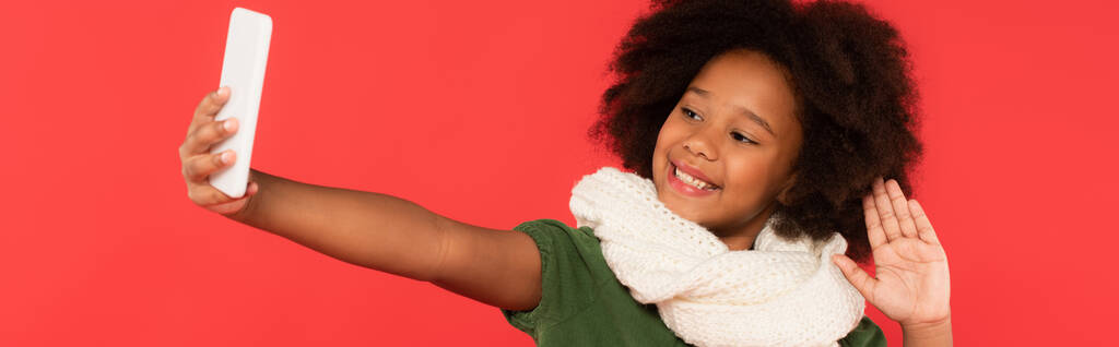 Enfant afro-américain souriant dans une écharpe chaude ayant un appel vidéo sur smartphone isolé sur rouge, bannière  - Photo, image