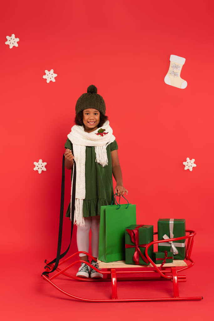Smiling african american child in scarf with mistletoe standing near sleigh, presents and shopping bag on red background - Photo, Image