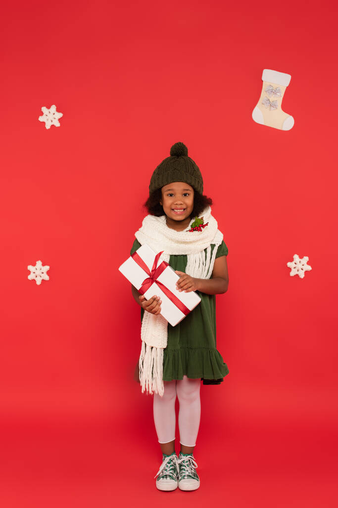 Felice ragazza afroamericana in maglia cappello e sciarpa con vischio tenere presente vicino a fiocchi di neve decorativi su sfondo rosso - Foto, immagini