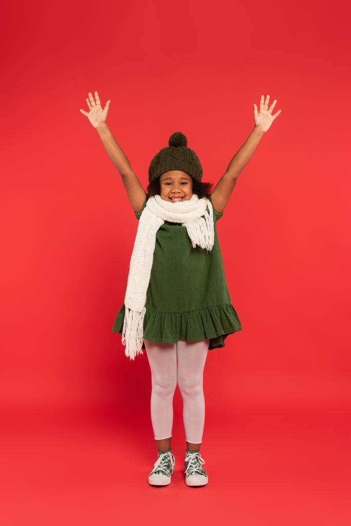 Afrikaans amerikaans kind met warme hoed en gebreide muts zwaaiend naar camera op rode achtergrond - Foto, afbeelding