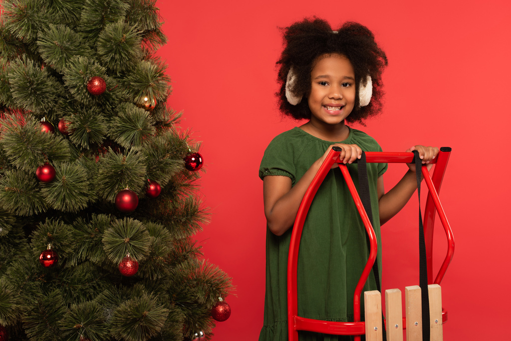 Mosolygó afro-amerikai gyerek fülvédőben áll közel szánkó és karácsonyfa elszigetelt piros  - Fotó, kép