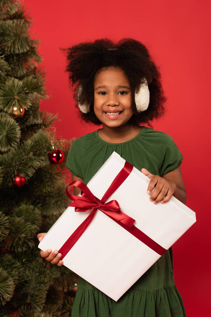 Ευτυχισμένο αφροαμερικανό παιδί σε ωτοασπίδες κρατώντας κουτί δώρου κοντά στο χριστουγεννιάτικο δέντρο απομονωμένο στο κόκκινο  - Φωτογραφία, εικόνα