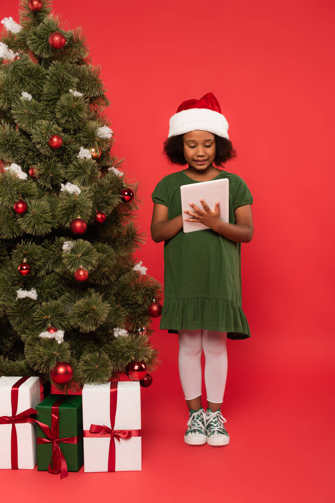 Noel Baba şapkalı gülümseyen Afrikalı Amerikalı çocuk hediye kutularının yanında dijital tablet ve kırmızı arka planda noel ağacı kullanıyor. - Fotoğraf, Görsel