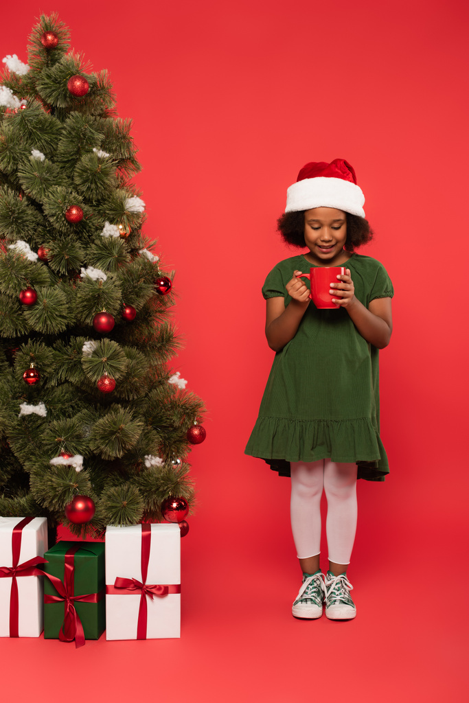 Afro-americana chica en santa hat celebración taza cerca de árbol de Navidad y cajas de regalo sobre fondo rojo - Foto, Imagen
