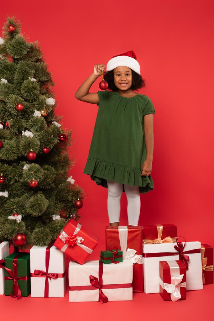 Usmívající se africké americké dítě v Santa klobouk drží vánoční míč v blízkosti dárky a jedle na červeném pozadí - Fotografie, Obrázek