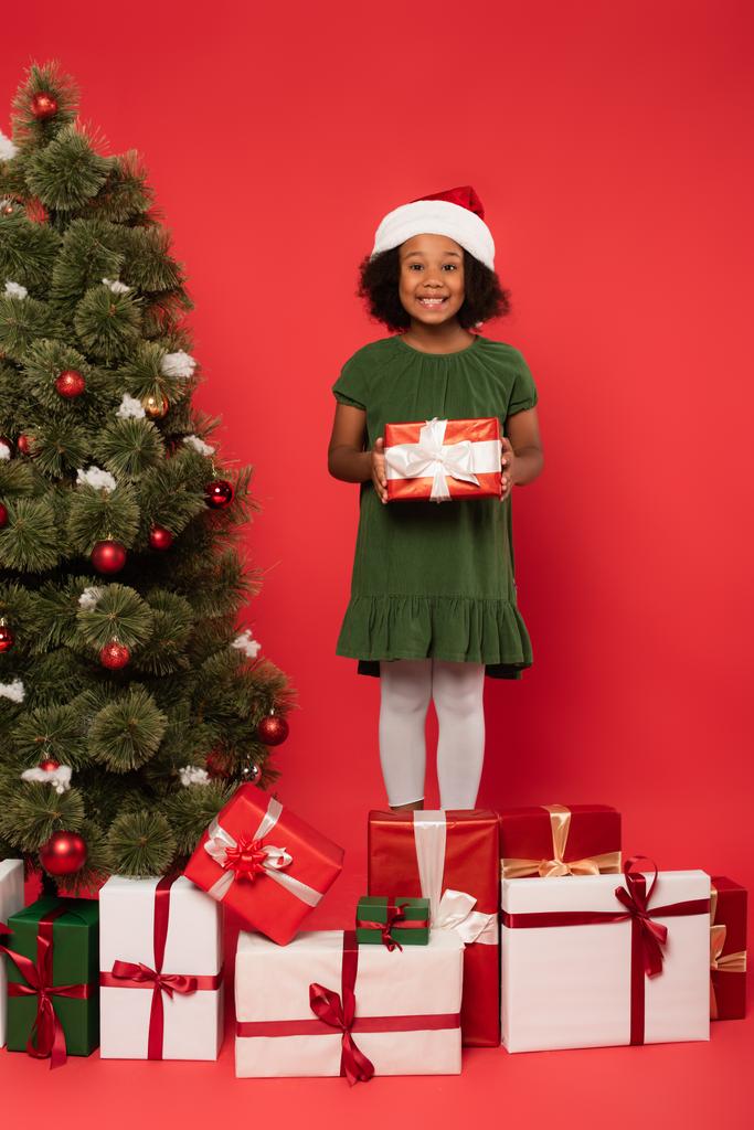 Bambino afroamericano in cappello da Babbo Natale in possesso di regalo vicino decorato albero di Natale su sfondo rosso - Foto, immagini