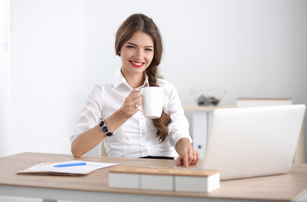 Junge Geschäftsfrau sitzt mit Tasse im Büro auf dem Schreibtisch - Foto, Bild