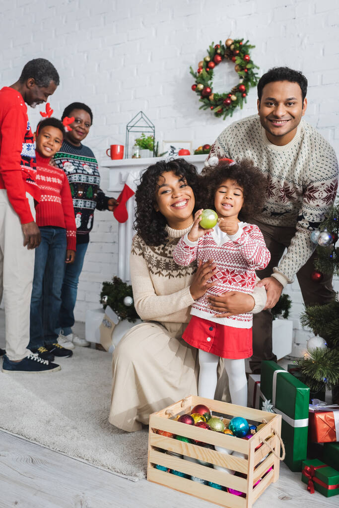 афроамериканська дівчина тримає різдвяний м'яч біля дерев'яної коробки з вафлями і щасливою сім'єю у вітальні
 - Фото, зображення