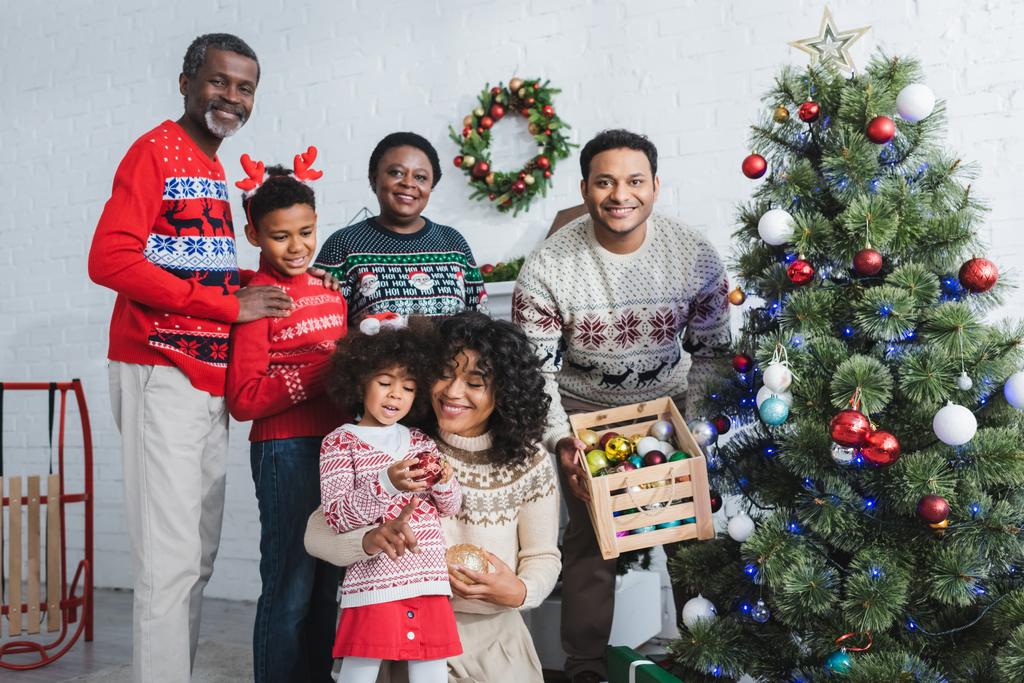 hombre sosteniendo caja de madera con bolas decorativas cerca del árbol de Navidad y la familia afroamericana - Foto, Imagen