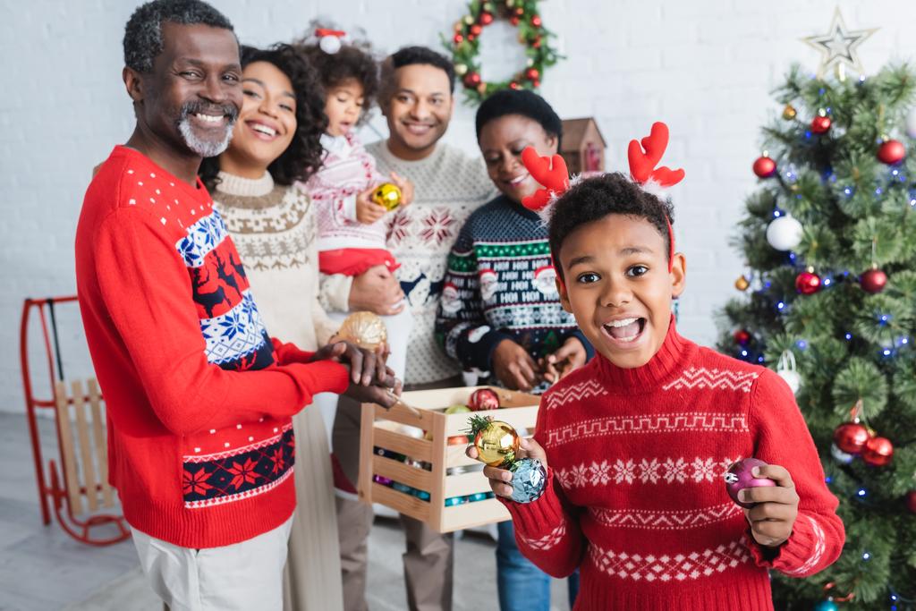 aufgeregten afrikanisch-amerikanischen Jungen in Rentierhörnern Stirnband hält Weihnachtskugeln in der Nähe glücklicher Familie - Foto, Bild