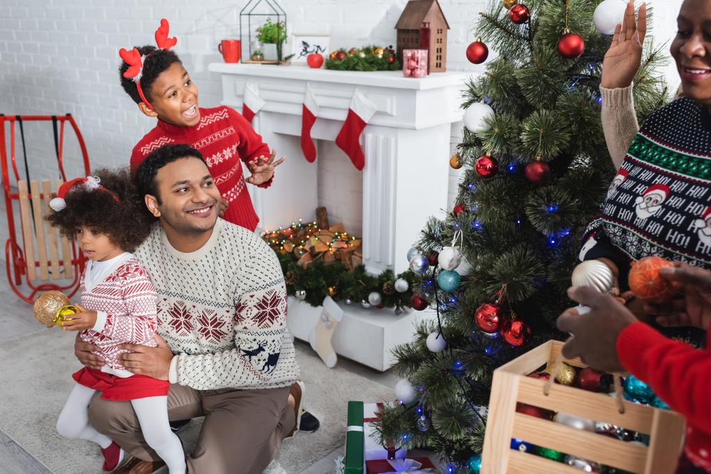 vzrušený africký americký chlapec mává rukou rozmazané prarodiče zdobení vánoční stromeček v blízkosti muže s dcerou - Fotografie, Obrázek