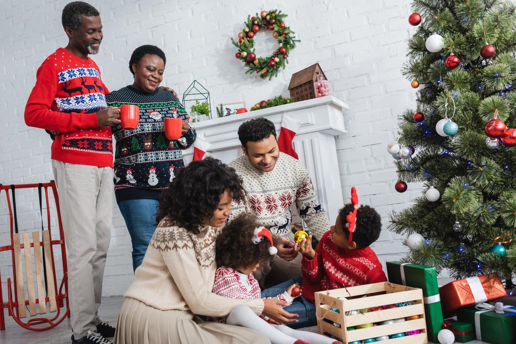 Afrikanisch-amerikanische Eltern mit Kindern wählen dekorative Christbaumkugeln in der Nähe des Weihnachtsbaums und Großeltern mit Tassen - Foto, Bild