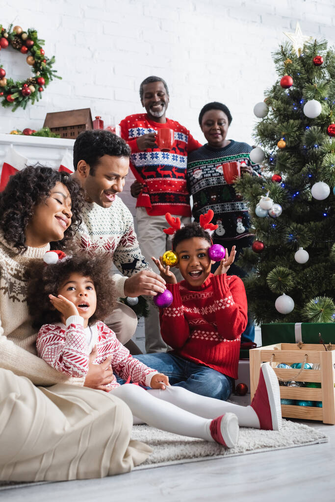 niño excitado en cuernos de reno diadema con adornos decorativos cerca del árbol de Navidad y sonriente familia afroamericana - Foto, imagen