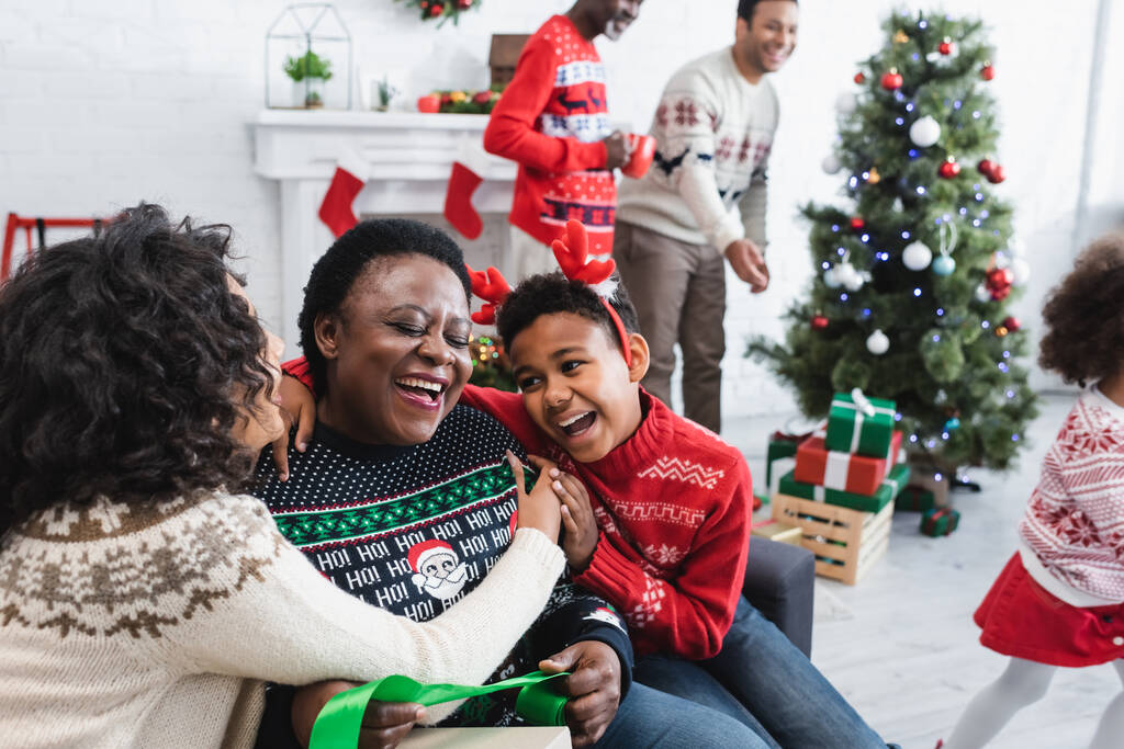 onnellinen poika ja nainen syleilee naurava afrikkalainen mummo lähellä hämärtynyt miesten ja joulukuusi olohuoneessa - Valokuva, kuva