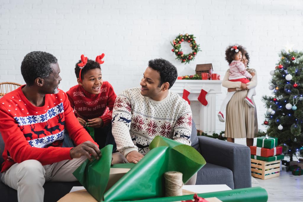 アフリカ系アメリカ人男の子でトナカイ角頭バンド笑顔近く男性パッキングプレゼントとぼやけた母親とともに子供クリスマスツリーの近く - 写真・画像