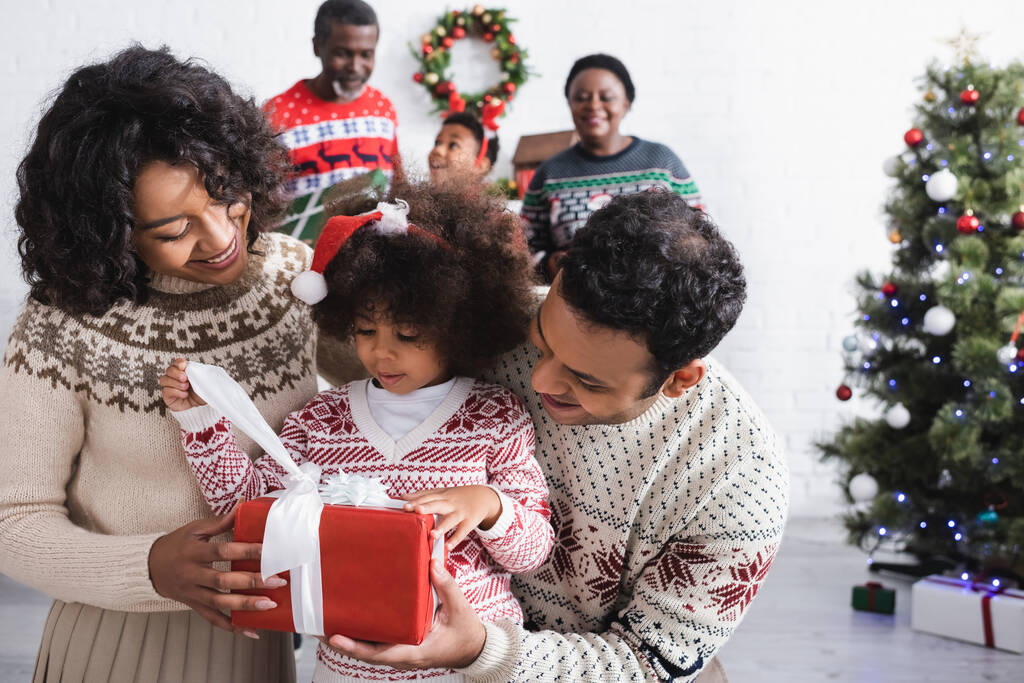 afro-amerikai lány télapó kalap gazdaság ajándék közelében boldog szülők és elmosódott család a nappaliban karácsonyfa - Fotó, kép