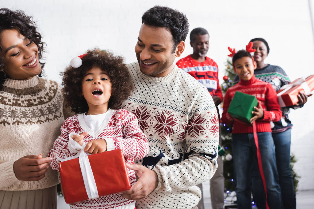 padres felices cerca de hija sorprendida con caja de regalo y familia afroamericana sobre fondo borroso - Foto, Imagen