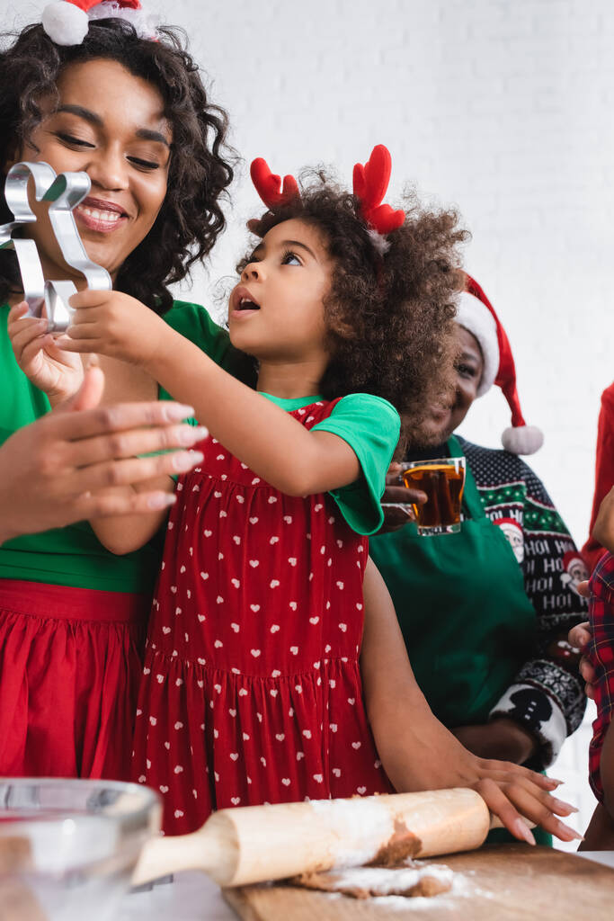 african american girl in reindeer horns headband holding cookie cutter near happy mom and smiling granny - Photo, Image