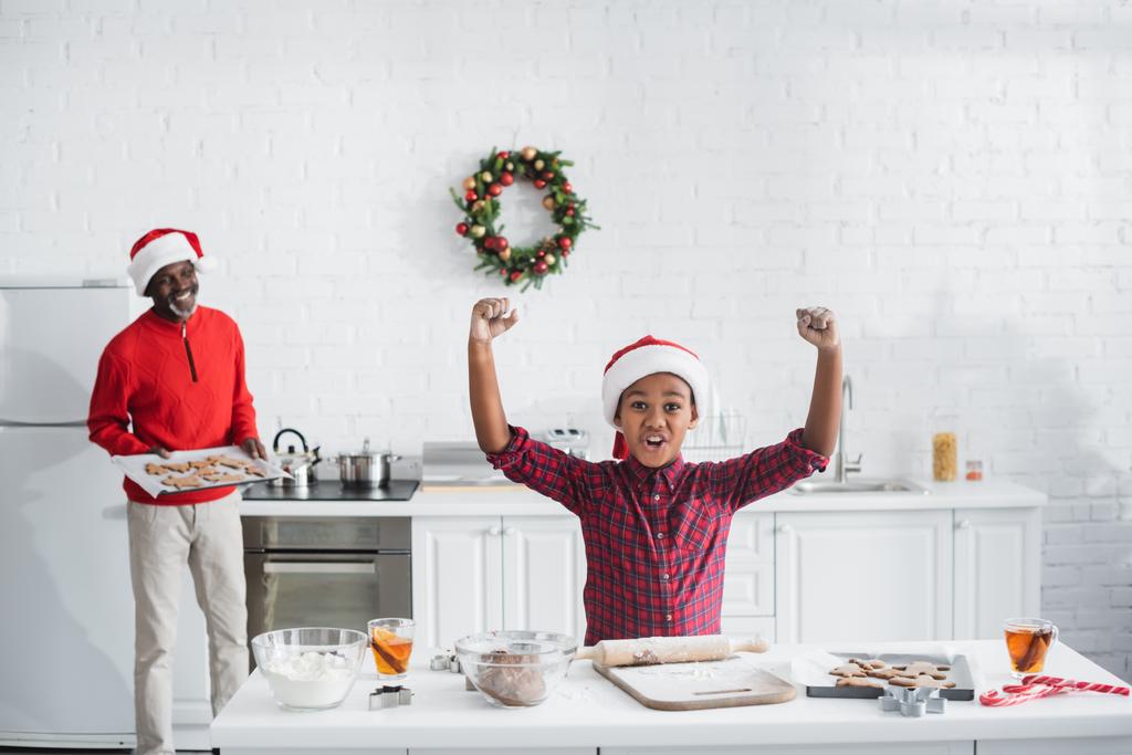 возбужденный африканский американец в шляпе Санты, показывающий успех жеста вблизи размытого деда с сырым рождественским печеньем на кухне - Фото, изображение
