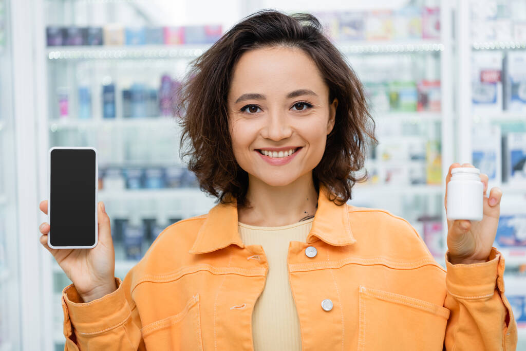 mujer alegre sosteniendo la botella con la medicación y el smartphone con la pantalla en blanco en la farmacia - Foto, Imagen