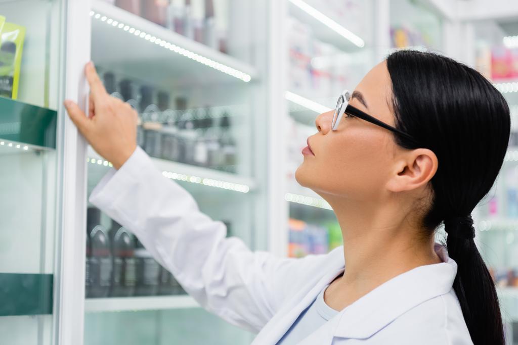 vista lateral do asiático farmacêutico em óculos olhando para medicação em prateleiras borradas - Foto, Imagem