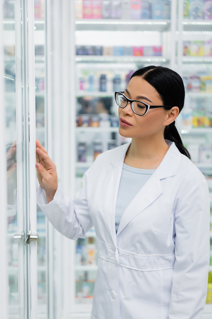 azjatycki farmaceuta w okularach patrząc na leki na rozmytych półkach - Zdjęcie, obraz