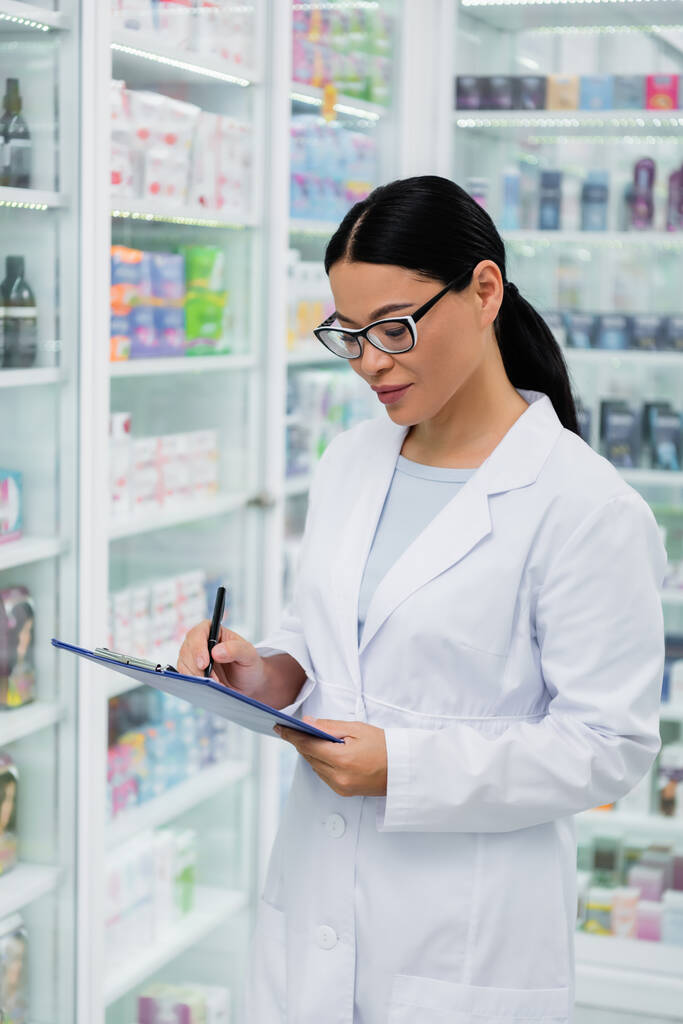 Азиатский фармацевт в очках, пишущий на планшете в аптеке  - Фото, изображение