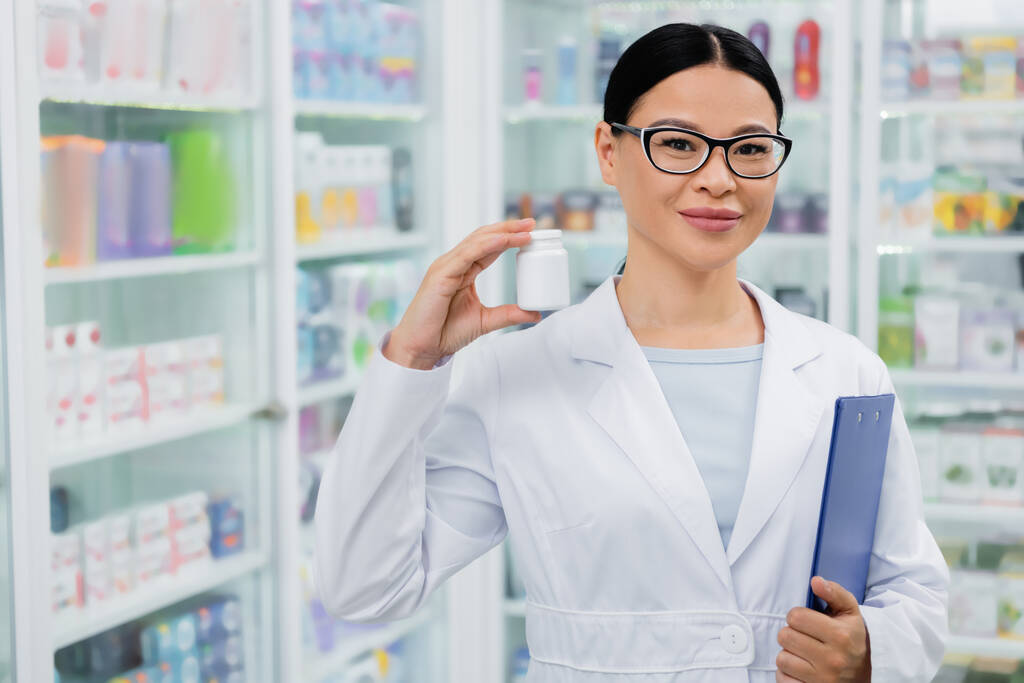 asiatischer Apotheker in Brille lächelt, während er Flasche mit Medikamenten und Klemmbrett in der Hand hält - Foto, Bild