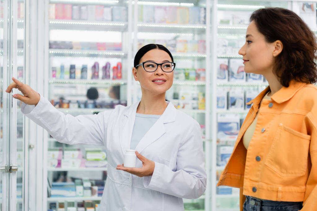 iloinen aasialainen farmaseutti lasit tilalla pullo huumeita ja osoittaa pois lähellä brunette asiakas apteekissa  - Valokuva, kuva