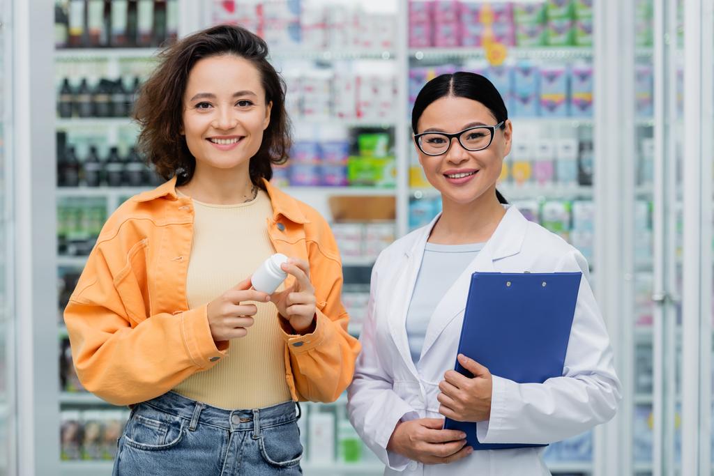 asiatischer Apotheker in Brille hält Klemmbrett in der Nähe einer fröhlichen Kundin mit Flasche in der Drogerie  - Foto, Bild
