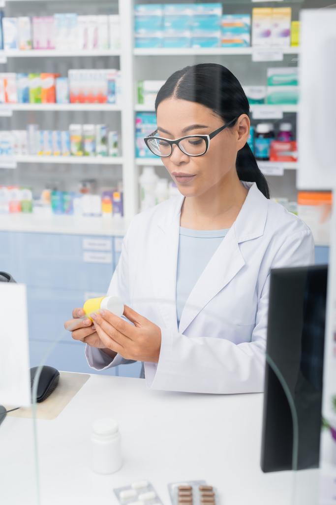 brunette asian pharmacist in glasses and white coat looking at bottle with medication at counter  - Photo, Image