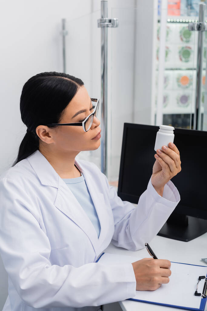 farmacéutico asiático en gafas y blanco abrigo mirando en botella con pastillas mientras escribe en portapapeles  - Foto, Imagen
