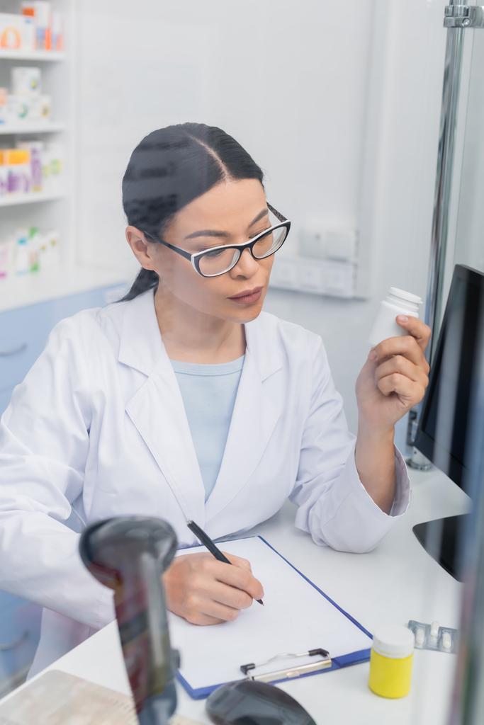 asian pharmacist in glasses holding bottle with medication while writing on clipboard in drugstore - Photo, Image