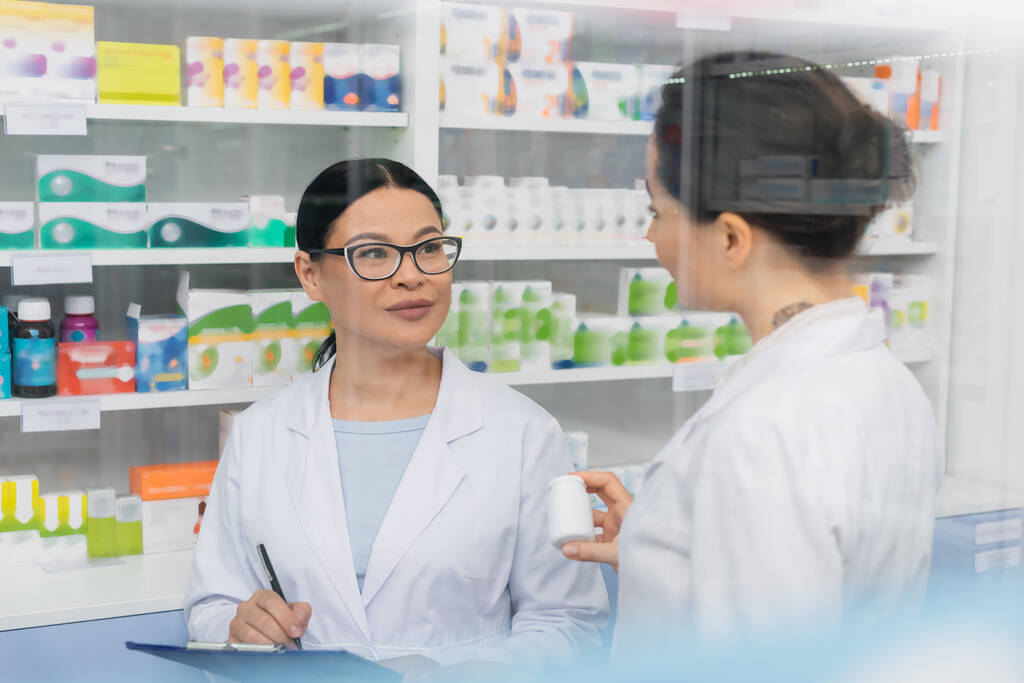 farmaceuta wytatuowany w białym fartuchu trzymający butelkę w pobliżu azjatyckiego kolegi w okularach piszących na schowku w aptece  - Zdjęcie, obraz