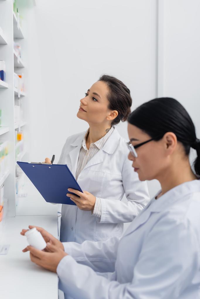白衣を着た薬剤師がクリップボードを持ちながら眼鏡でアジア人の同僚のぼやけた薬をチェック  - 写真・画像