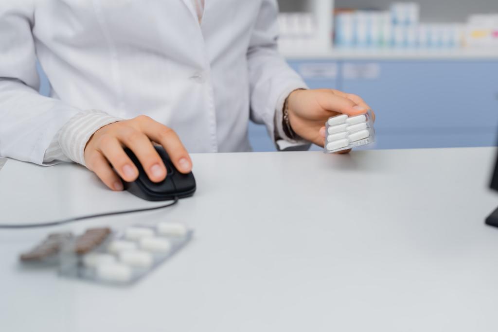 обрізаний вид фармацевта, який тримає блістерний пакет і використовує комп'ютерну мишу в аптеці
 - Фото, зображення