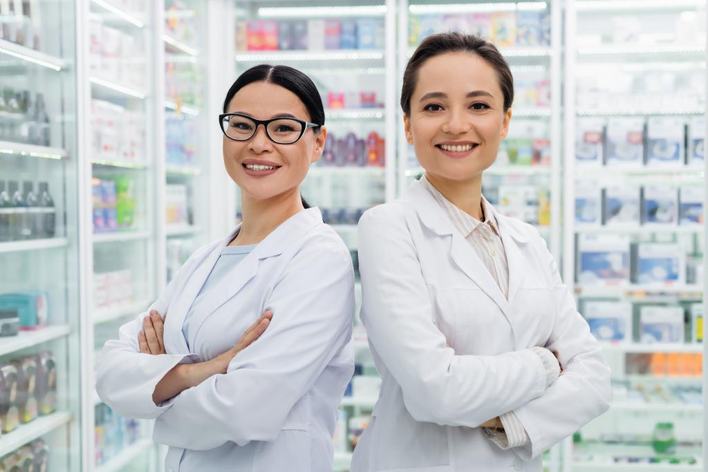 Χαρούμενοι διαφυλετικοί φαρμακοποιοί με λευκά παλτά που στέκονται με σταυρωμένα χέρια στο φαρμακείο - Φωτογραφία, εικόνα