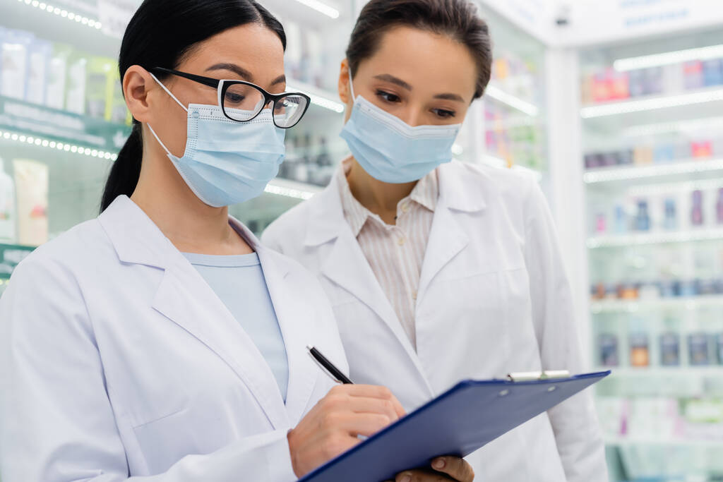 азіатський фармацевт в медичній масці пише в буфері біля колеги
  - Фото, зображення