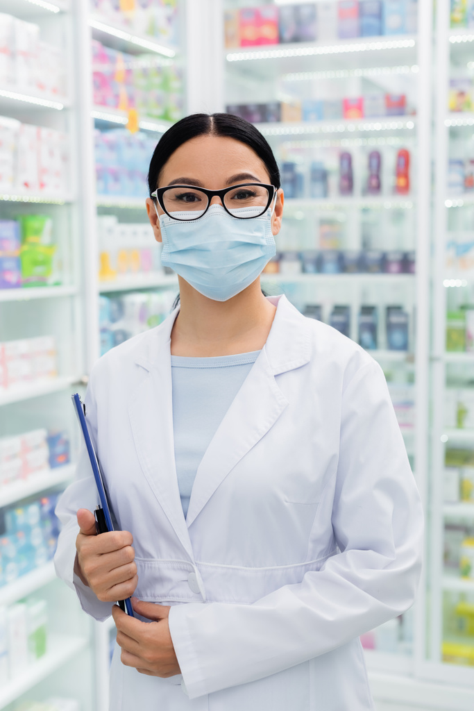 azjatycki farmaceuta w okularach i masce medycznej trzymając schowek w aptece  - Zdjęcie, obraz