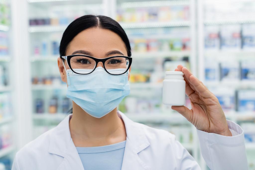 asiatique pharmacien dans lunettes et masque médical tenant bouteille avec des pilules - Photo, image