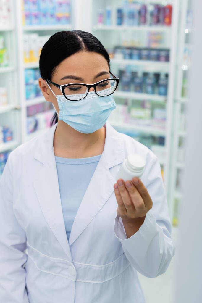 Азиатский фармацевт в очках и медицинской маске смотрит на бутылку с таблетками - Фото, изображение