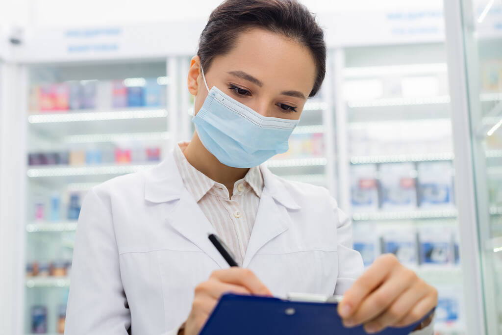 brunette apothecary in medical mask writing on clipboard  - Photo, Image